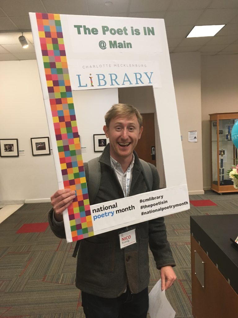 Nico, a white man with blonde hair, holding up a poster board with a square hole cut out and smiling through the hole. The poster board says "Charlotte Mecklenburg Library National Poetry Month" on the edges around his face.